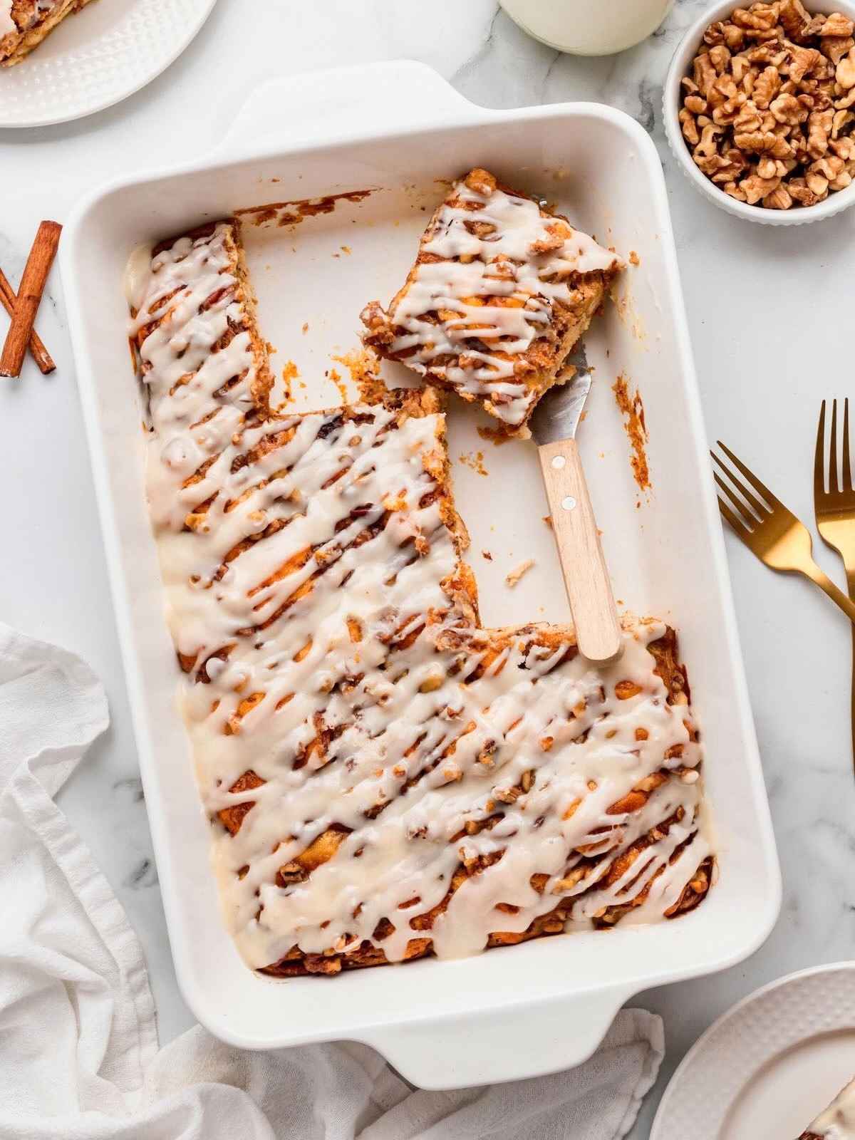 Iced pumpkin cinnamon roll cake in a white baking dish with a spatula, some pieces served, walnuts on the side.