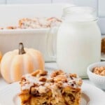 A slice of glazed pumpkin cinnamon roll casserole with walnuts on a plate, next to a jar of milk and a small white pumpkin.