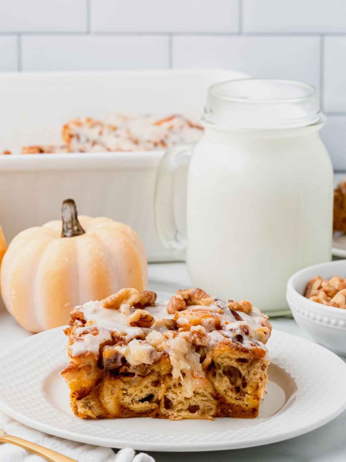 A slice of glazed pumpkin cinnamon roll casserole with walnuts on a plate, next to a jar of milk and a small white pumpkin.