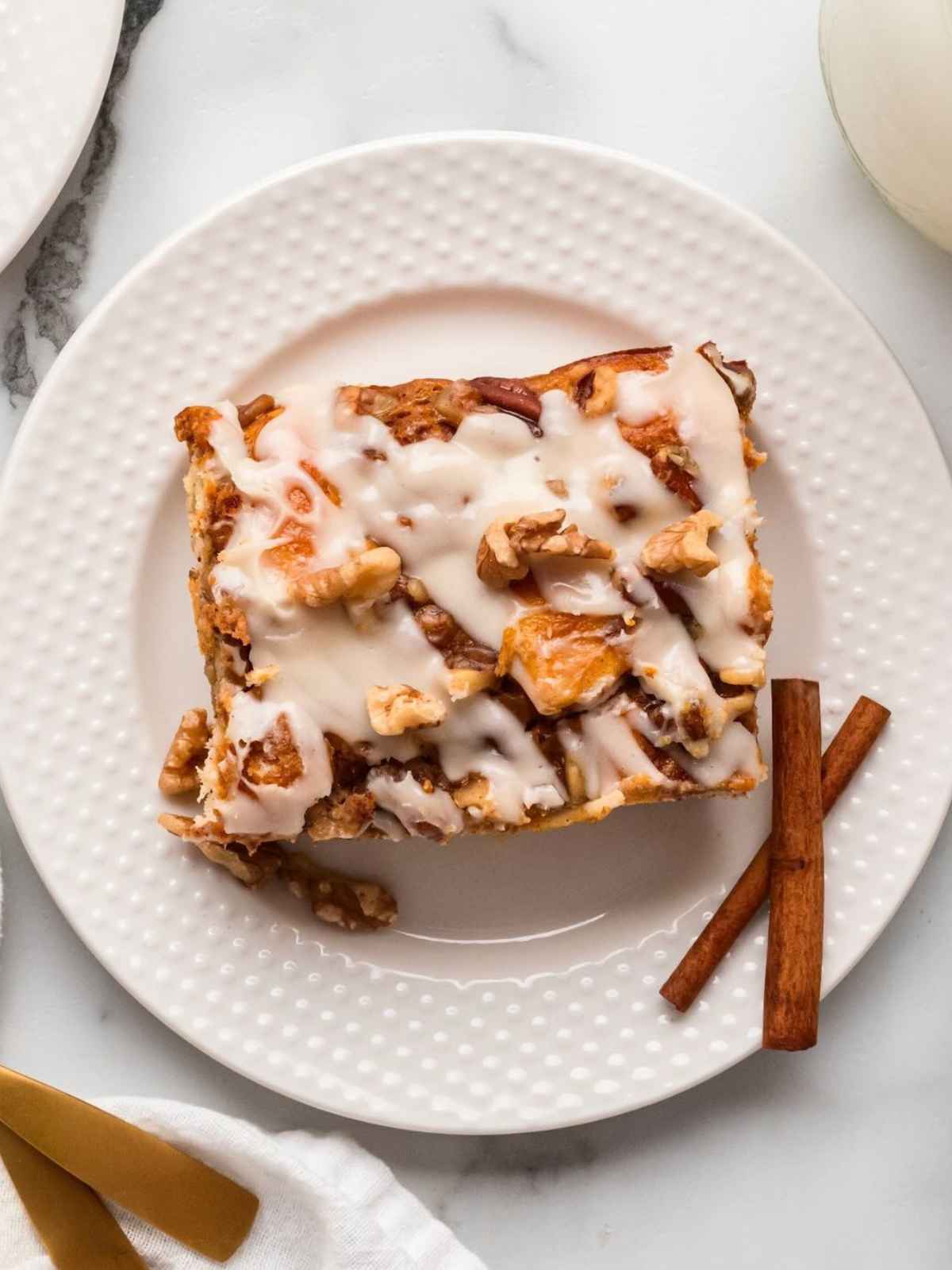 Iced cinnamon roll with walnuts on a white plate beside two cinnamon sticks, viewed from above.