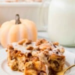 A slice of pumpkin casserole with walnuts and icing on a plate, with a fork, milk, and a small pumpkin nearby.