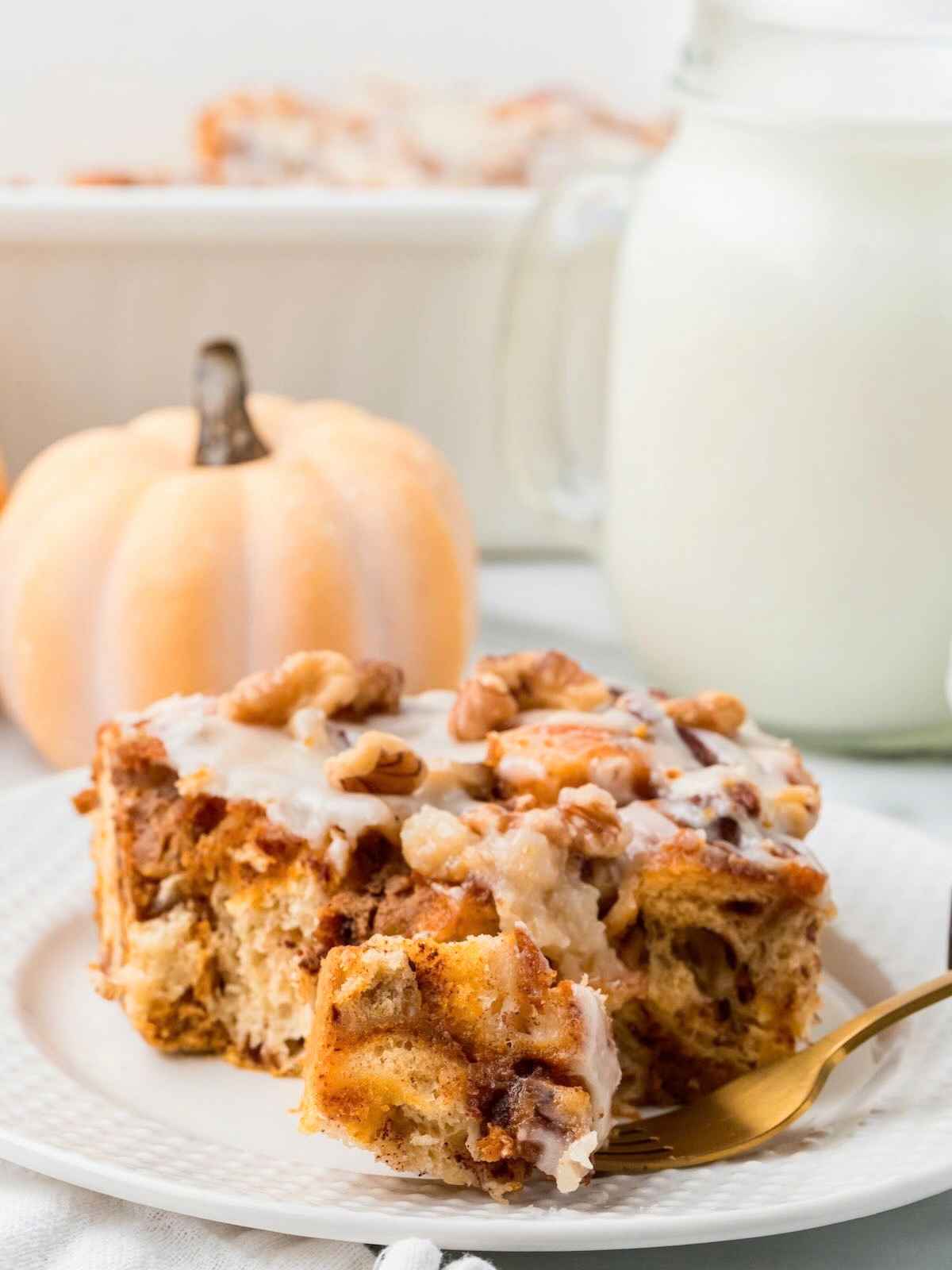 A slice of pumpkin casserole with walnuts and icing on a plate, with a fork, milk, and a small pumpkin nearby.