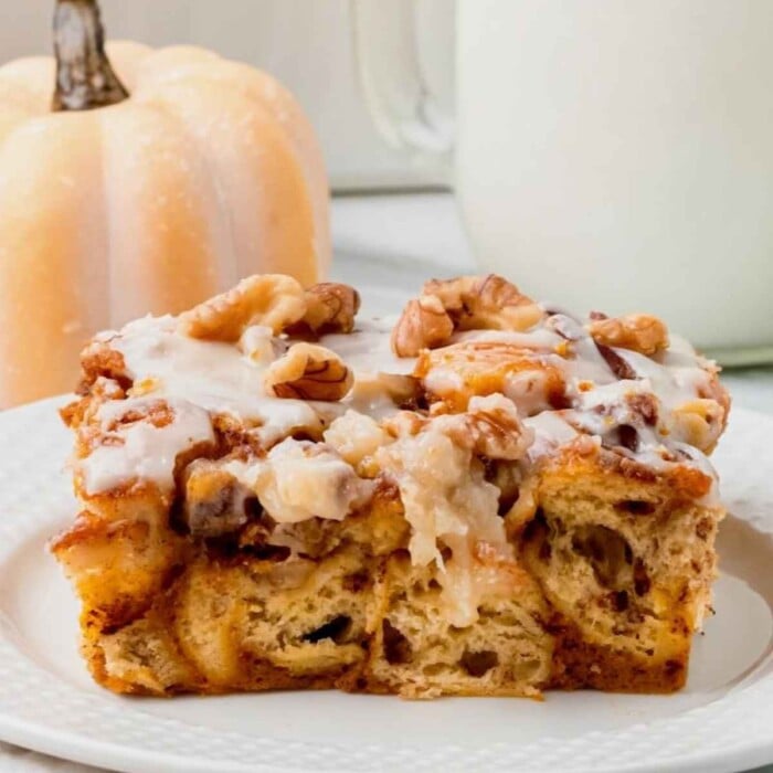 A slice of glazed pumpkin cinnamon roll casserole with walnuts on a white plate; small pumpkin in background.