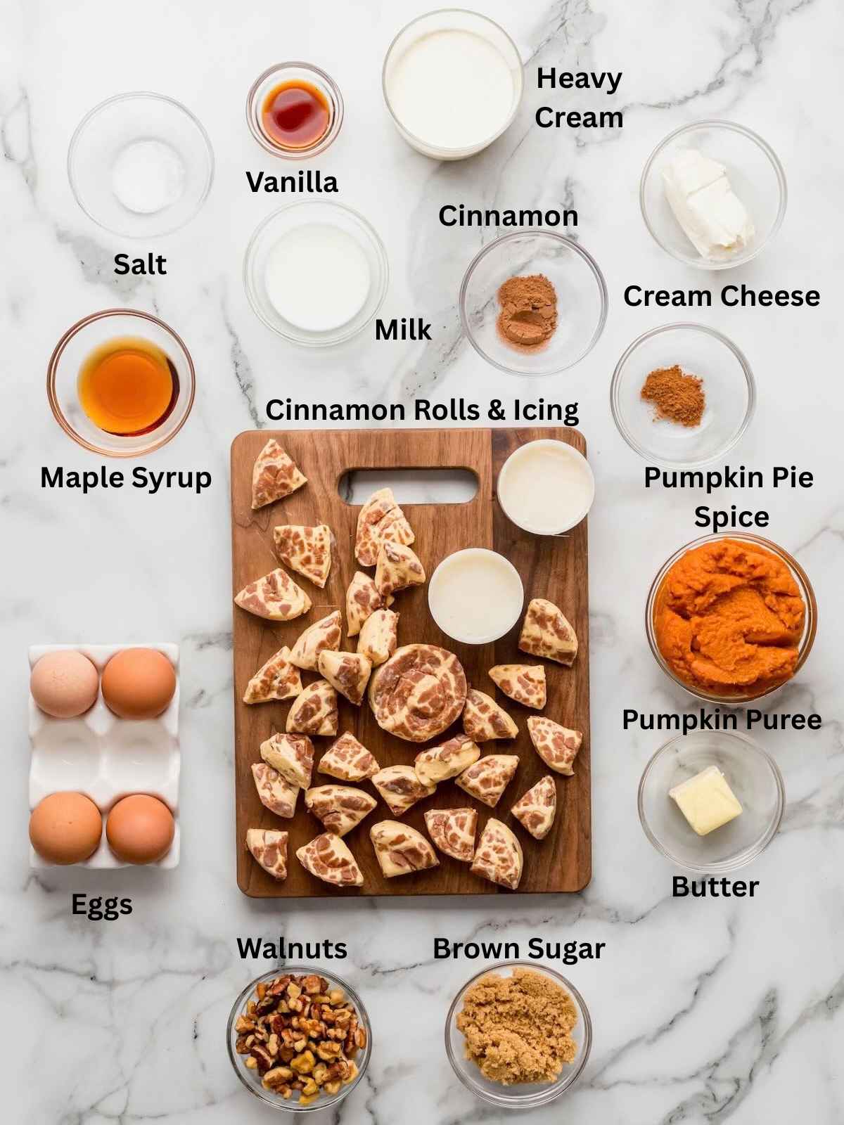 Ingredients for cinnamon roll casserole, labeled and arranged on a marble surface with a wooden board in the center.