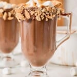 Glass mug of hot chocolate with a smores-inspired rim of melted chocolate, graham cracker crumbs, and marshmallows.