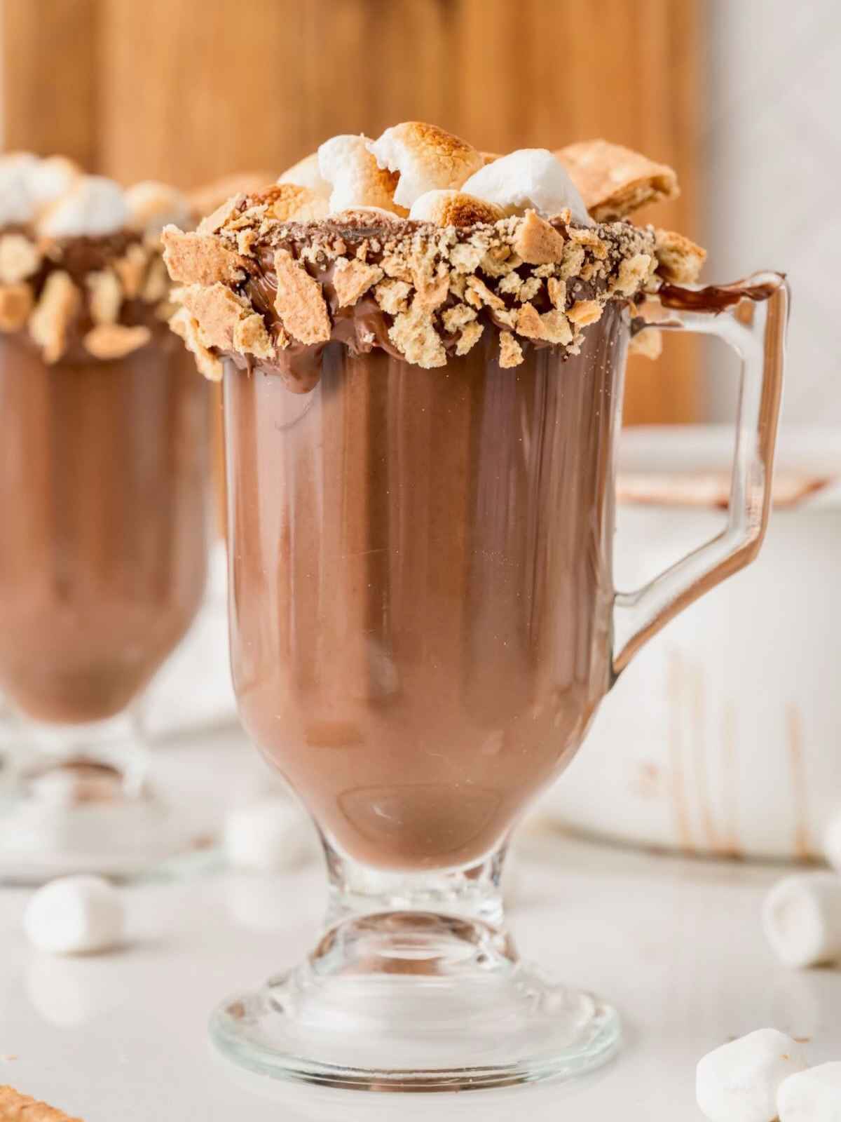 Glass mug of hot chocolate with a smores-inspired rim of melted chocolate, graham cracker crumbs, and marshmallows.