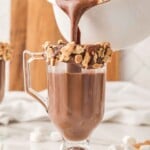 Hot chocolate being poured into a glass mug rimmed with chocolate and crushed graham crackers.