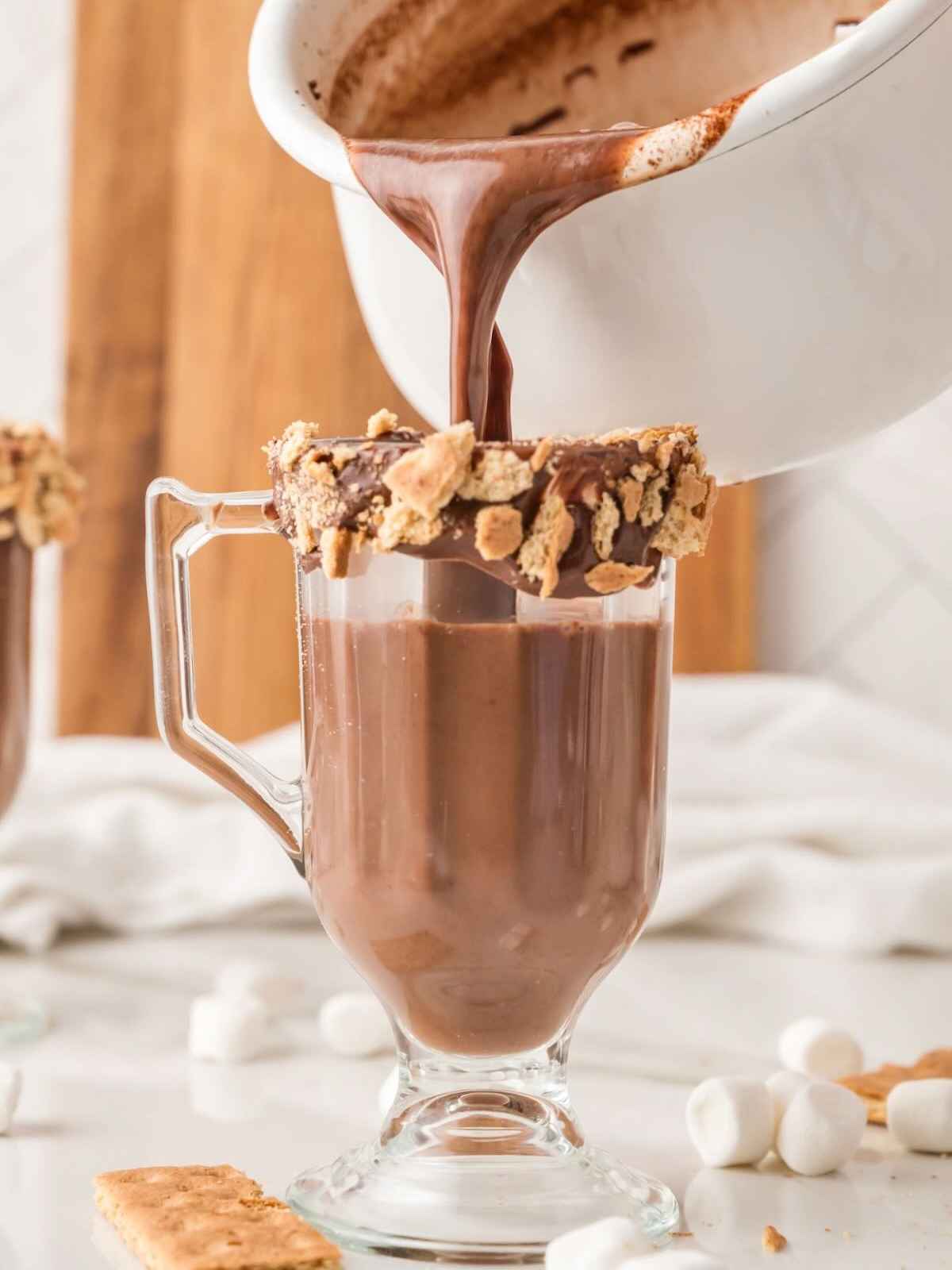 Hot chocolate being poured into a glass mug rimmed with chocolate and crushed graham crackers.