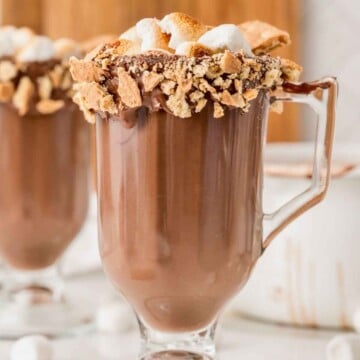 Glass mug of hot chocolate topped with toasted marshmallows and a graham cracker rim.
