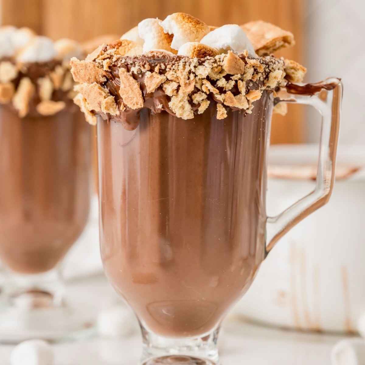 Glass mug of hot chocolate topped with toasted marshmallows and a graham cracker rim.